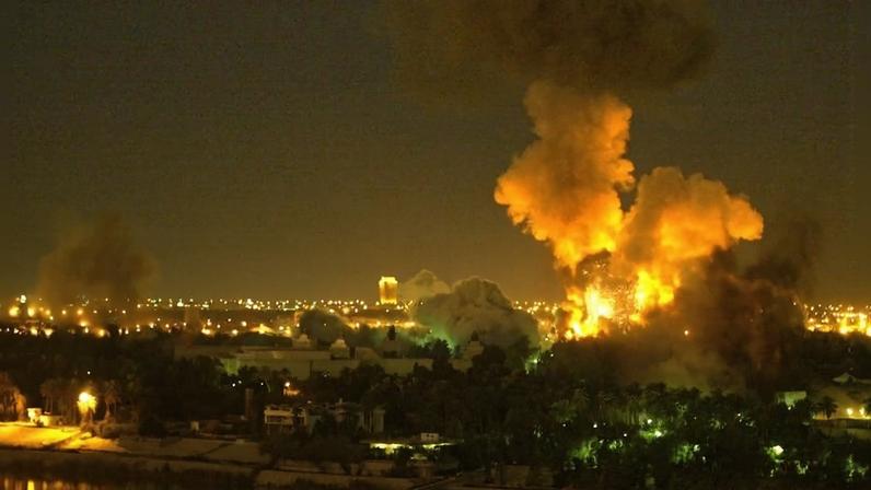 Smoke and yellow flames billow from a building in the foreground in this wide shot showing the city skyline of Baghdad at night. Other smaller plumes of smoke rise from buildings nearby, merging into a general haze over the sky, obscuring all stars.