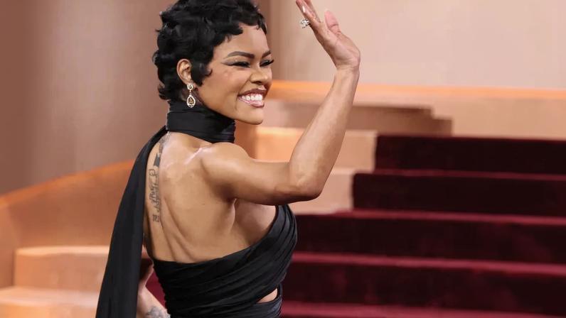 Teyana Taylor in a black gown with a cut-out midriff and diamond-studded bow, waving to the camera.