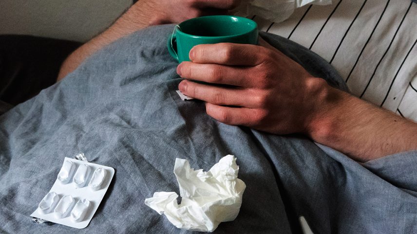 Fotografia de um homem deitado na cama com lenços, xícara de chá, spray nasal e comprimidos.