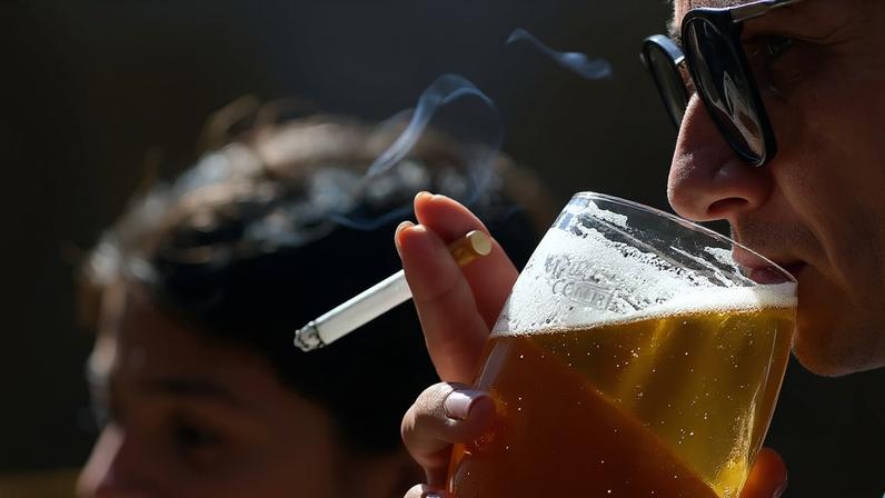 Fotografia de uma pessoa com um cigarro na mão bebe uma cerveja.