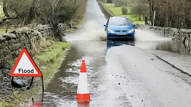 BBC A blue car driving through a small flood in a country road. There is a hazard sign warning of the flood and a cone in some standing water.