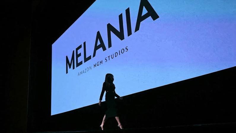 A silhouette of Melania Trump appears a she walks in front of a giant movie screen showing the title of her new movie, Melania.
