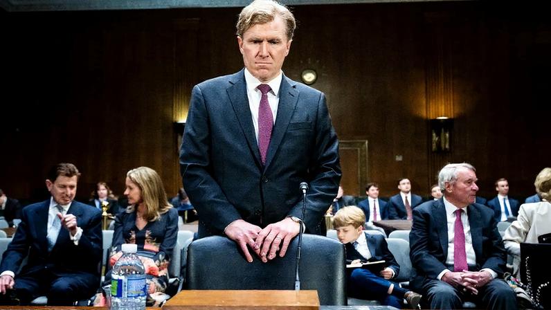 Elbridge Colby stands behind a chair with a microphone, about to sit down. He wars a dark business suit and a serious expression. Behind him are seated officials, aides, and one child.