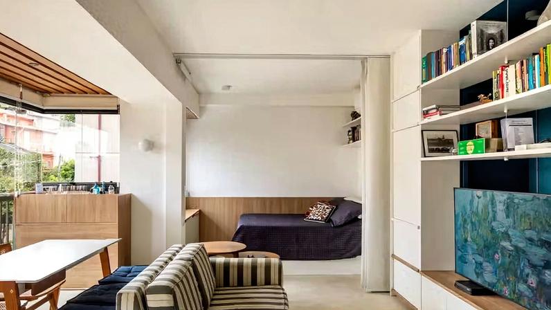 LIVING | O apartamento é totalmente integrado, inclusive com o quarto conectado ao estar, porém podendo ser isolado pela cortina deslizante da Hunter Douglas. A marcenaria é um item importante no projeto, com desenho do escritório, para que os móveis se concentrem nas paredes, liberando o máximo espaço possível para o uso, como a estante, a meia-parede da cama e o bar na varanda, executados em MDF laminado pela Casttini e montada pela Dell Ambiente Studio