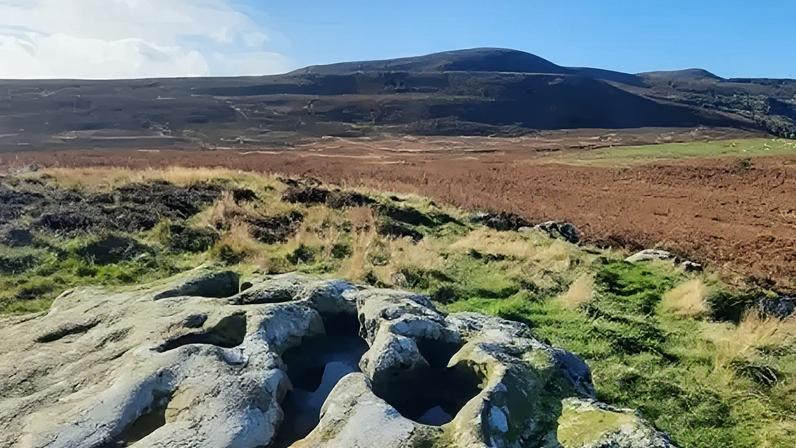 Duncan Hutt A landscape photo of an area of the Rothbury Estate. The picture has been taken from the top of a large boulder, which can be seen in the foreground. It looks out over grassland and the Simonside Hills can be seen in the distance. They are dark, almost black, in colour with rounded peaks above a high ridge.