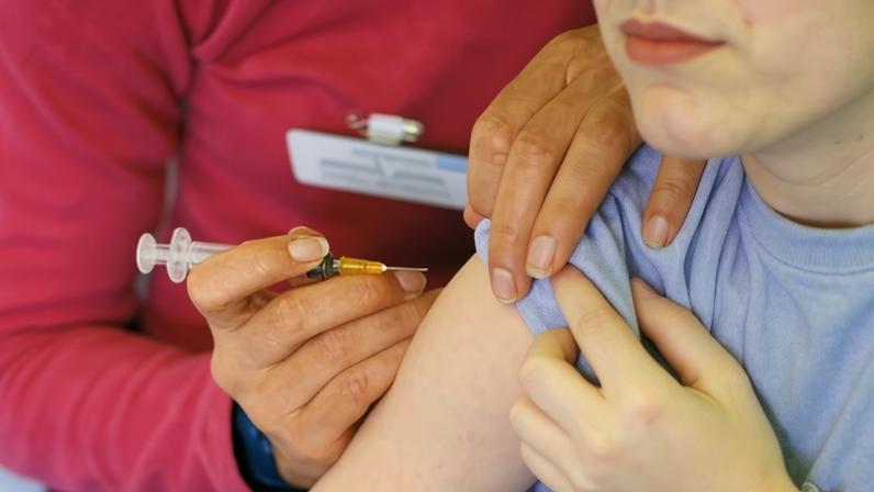 A child is about to be given the MMR (mumps, measles, rubella) vaccination by a surgery nurse. The nurse is holding the girl's sleeve up over her shoulder and bringing a syringe to her skin.