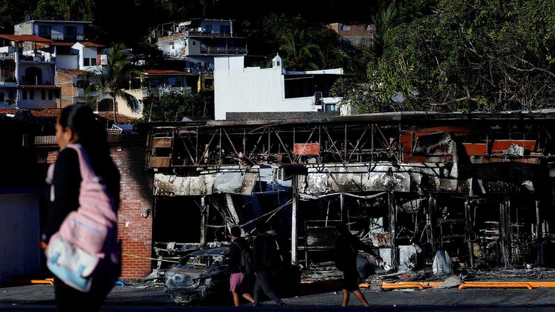 People walk past a store and a car that were set on fire by members of organized crime, in the aftermath of a Mexican military operation in which Jalisco New Generation Cartel (CJNG) leader Nemesio Oseguera, known as "El Mencho," was killed, in Puerto Vallarta, Mexico, February 26, 2026. REUTERS/Daniel Becerril
