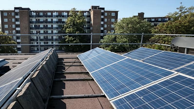 Solar panels are installed in rows on the roof of a block of flats in Brixton, London. Another block is seen in the background against a clear blue sky.