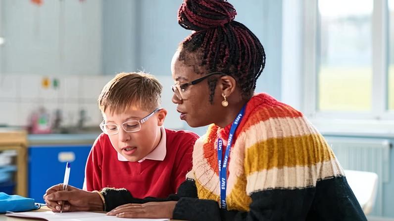 A Send teacher in glasses and a striped jumper sits at a school desk helping a student with special educational needs