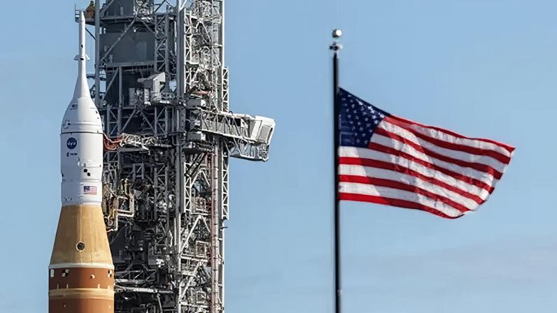 EPA/ Shutterstock Orange and white Artemis rocket next to a towering grey space system and a blue, white, and red starts and stripes flag, against a blue sky