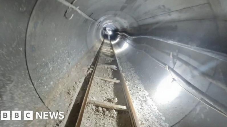 A view down an underground tunnel that is lit with bright white artificial light. There is a ladder track on the floor and various pipes and wires along the concrete walls.