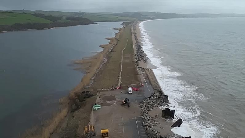 A drone view of the A379 Slapton Line between Torcross and Slapton. It shows the sea on one side and a lake on the other side. The road is damaged in two parts where maintenance vans can be seen.