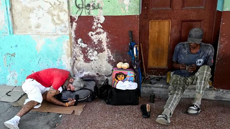 Two homeless men sit against a wall on a street in Cuba. One man is perched on a step, looking at his hands, while the other curls up to lie down while using a duffel bag as a pillow. A few possessions, including umbrellas, other bags, and a Dora the Explorer suitcase sit between them.