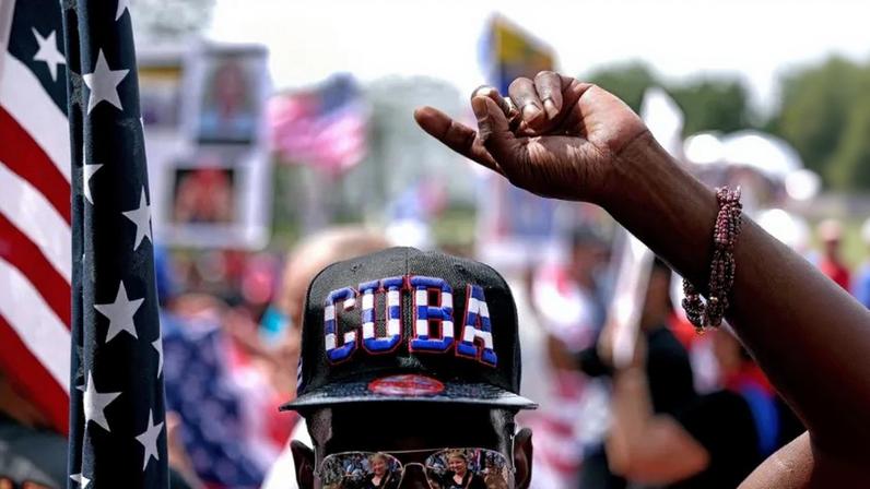 The top half of a man's face is shown—he's wearing aviator shades and a hat that reads CUBA. His hand is pointed in the air and he's next to a U.S. flag.