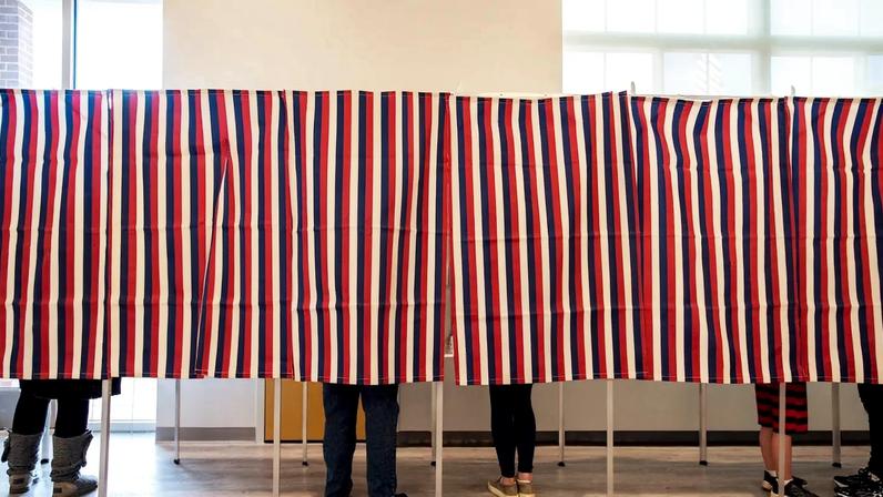 People stand inside inside booths that are partly covered by curtains in red, white, and blue stripes. Because of the curtains, only the voters' lower legs and feet are visible.
