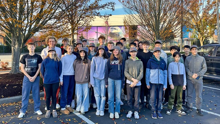 About 30 high school students and a teacher stand in front of a Boeing factory on a fall day
