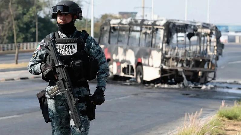 A Mexican National Guard member stands near a burnt bus set on fire.