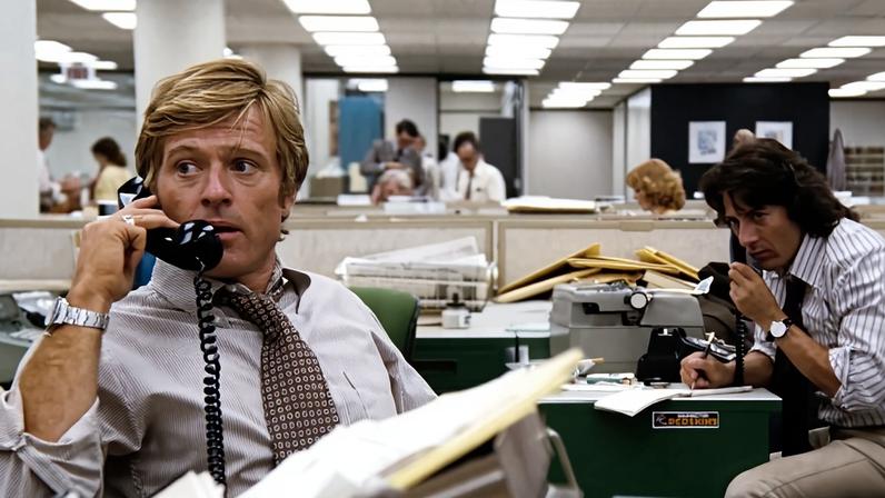 Robert Redford in a shirt and tie holds a phone to his ear as he sits behind a cluttered desk in an newsroom. Behind him, at right, Dustin Hoffman does the same, writing in a reporters notebook.