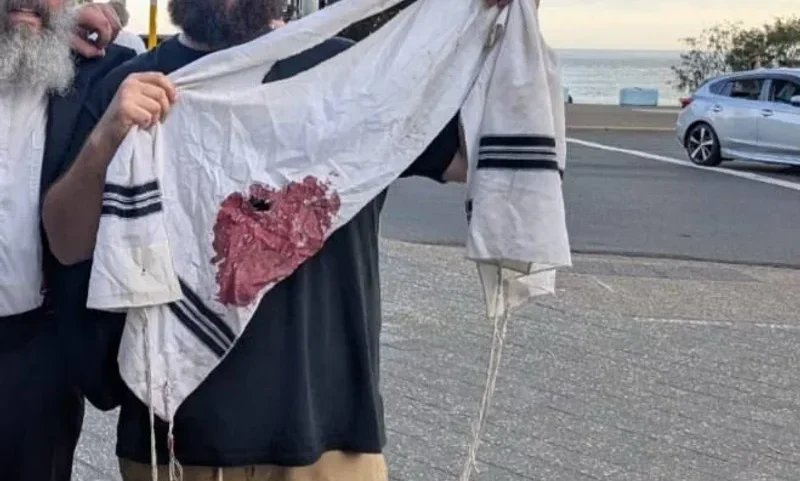 A imagem retrata dois homens segurando um talit manchado de sangue, simbolizando a tragédia ocorrida durante a celebração do Hanukkah em Sydney. O cenário é a praia de Bondi, com o mar ao fundo, sugerindo um contraste entre a beleza do local e a gravidade do evento. A expressão dos homens reflete dor e indignação, capturando a intensidade emocional do momento.