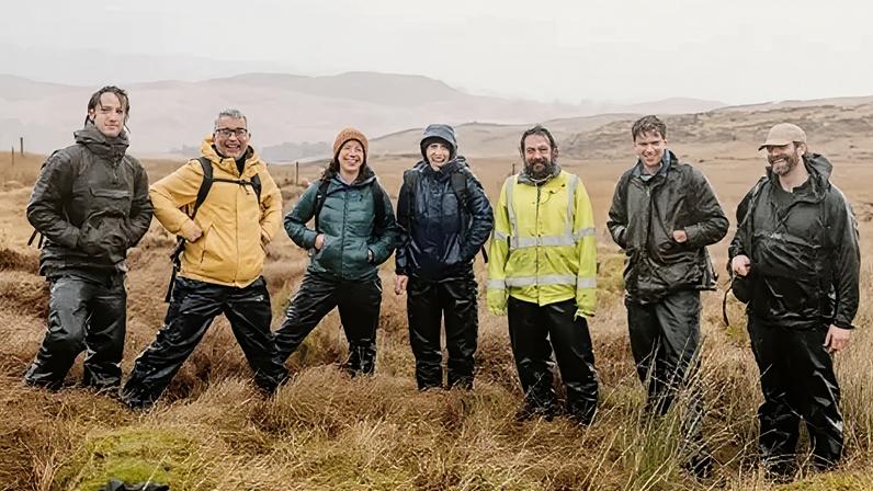 Five men and two woman wearing waterproof outerwear, smiling and standing on open grassland.