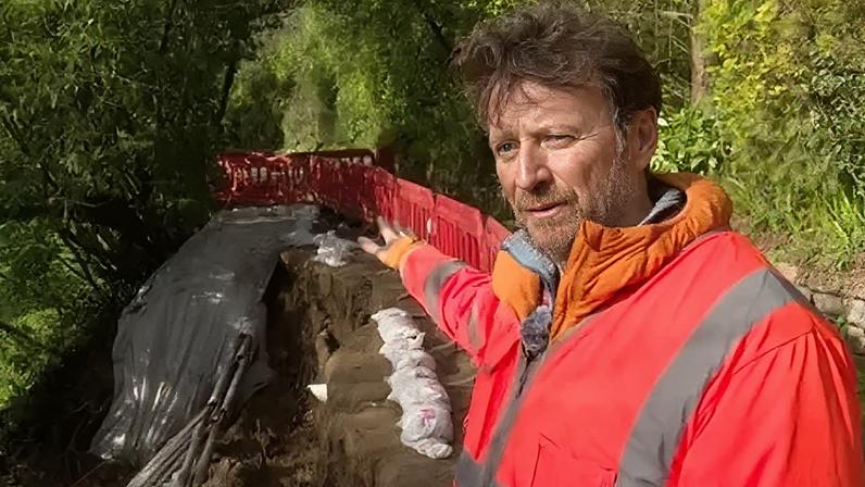 Tristen Dodd pictured on the Railway Walk. The landslip is visible behind him. He is gesturing towards it. He's wearing an orange hi-vis jacket. He has short brown hair and a beard.