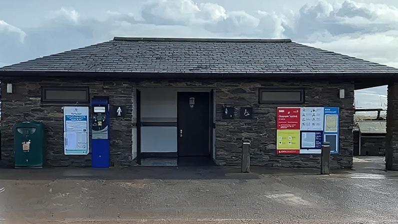 The public toilets at Torcross in Devon. The building is stone and there is a ladies sign to the left of the door. There is a parking machine next to the sign and on the other side is a sign.