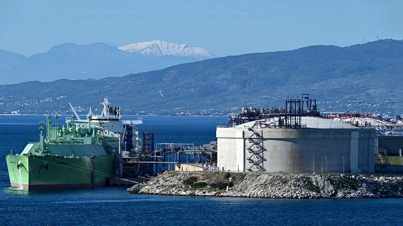 A tanker is shown in the water, adjacent to a terminal. There are mountains in the background.