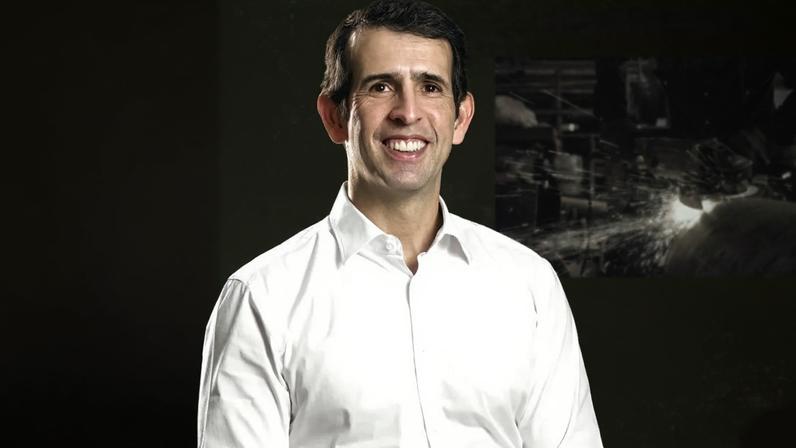 Homem sorrindo em P&B. Fundo roxo com elementos industriais: barras de metal e faíscas de esmerilhadeira.