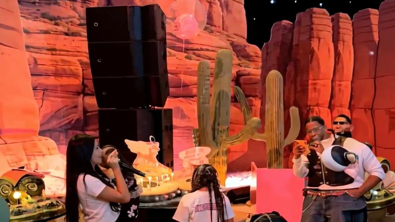 Kylie Jenner holding a microphone, standing next to a birthday cake, while Travis Scott stands nearby taking a picture with his phone.