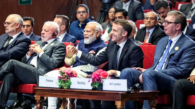 The four leaders sit in red upholstered chairs in the front row of an audience of other officials and aides. Modi and Macron smile and hold hands together, whereas Lulas has a serious expression and his hands clasped in front of him.