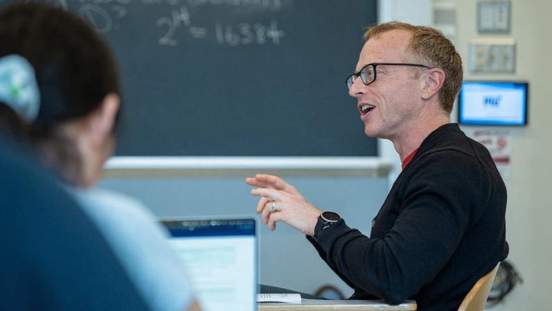 Brian Hedden sits at a desk in a classroom as he speaks to students seated around him.