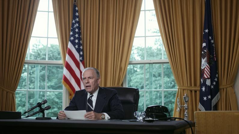 Gerald Ford, holding a piece of paper, speaks into microphones as he sits at the Resolute Desk in the Oval Office. Golden curtains on tall windows and flags on stands are seen behind him.