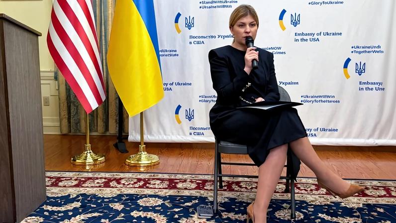 Olha Stefanishyna speaks into a microphone while sitting in front of U.S. and Ukrainian flags.