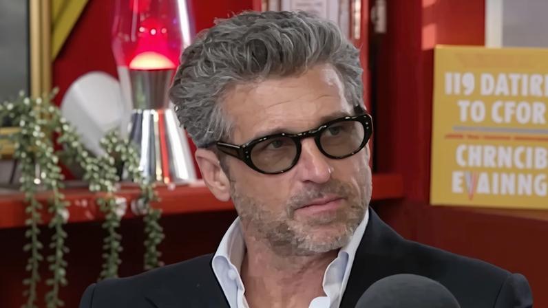Patrick Dempsey in a dark suit jacket, white shirt, and sunglasses, speaking into a Virgin Radio UK microphone.