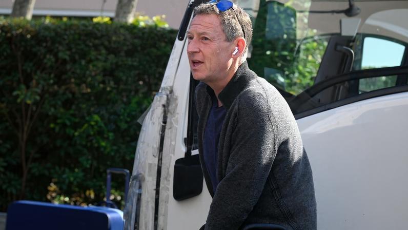 Michael Feldman walking with luggage near a white vehicle.