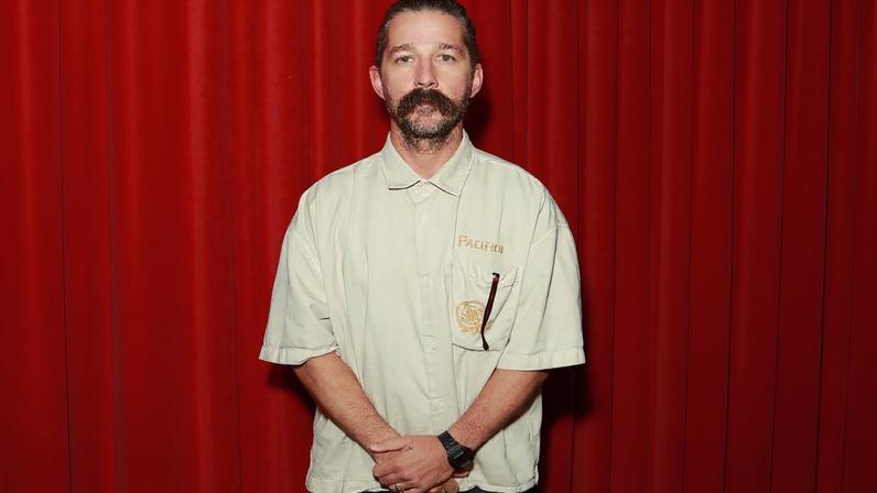 Shia LaBeouf stands with his hands clasped, wearing a light-colored button-up shirt and dark pants, against a red backdrop.