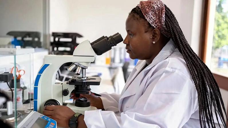 Fotografia de uma mulher negra cientista examinando espécimes sob microscópio em laboratório de patologia.