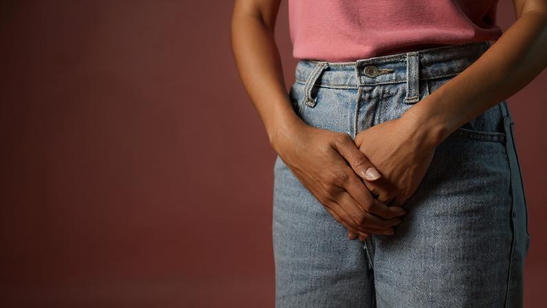 A imagem mostra a parte superior do corpo de uma pessoa, com as mãos cruzadas na frente do corpo. A pessoa está vestindo uma camiseta rosa e calças jeans claras. O fundo é de uma cor rosa suave, criando um contraste com a roupa. As mãos estão posicionadas de forma a cobrir a região do abdômen.