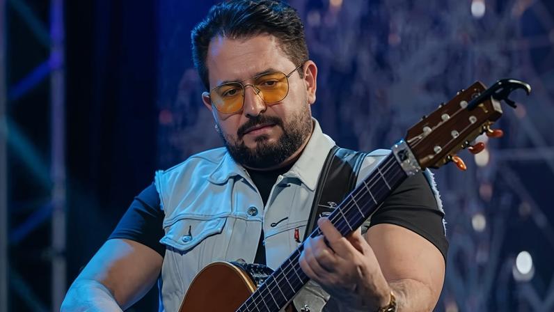 Um homem está tocando um violão em um palco. Ele usa uma camisa preta e um colete jeans. O homem tem barba e óculos escuros. O fundo é iluminado com luzes azuis e há uma estrutura metálica visível.