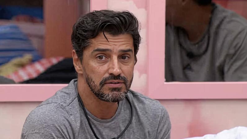 Homem de cabelos escuros e barba por fazer, veste camiseta cinza, sentado em ambiente interno com parede rosa ao fundo. Reflexo dele aparece em espelho atrás.