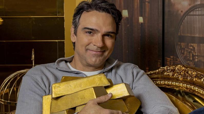 Homem de cabelos escuros e suéter cinza está sentado em cadeira dourada ornamentada, segurando várias barras de ouro empilhadas no colo, com fundo que sugere ambiente de cofre ou banco.