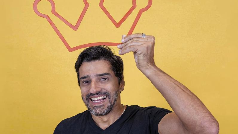 Homem de camiseta preta e calça jeans sorri enquanto segura uma coroa vermelha desenhada acima da cabeça, com fundo amarelo.