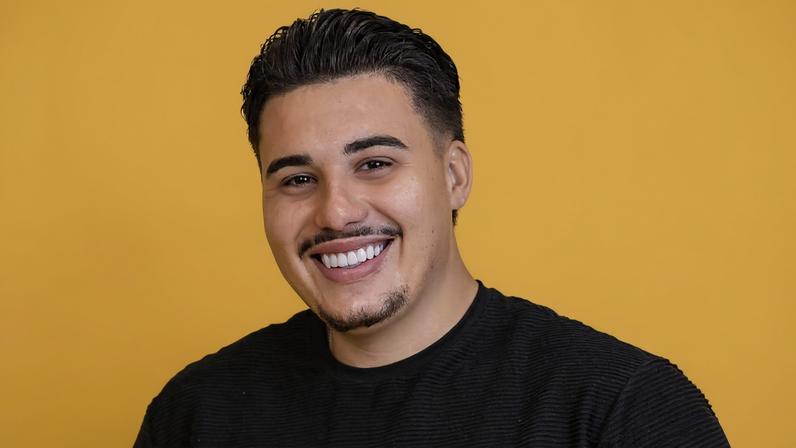 Homem jovem de cabelo curto e escuro sorri para a câmera, vestindo camiseta preta e calça bege, com as mãos nos bolsos, em fundo amarelo liso.
