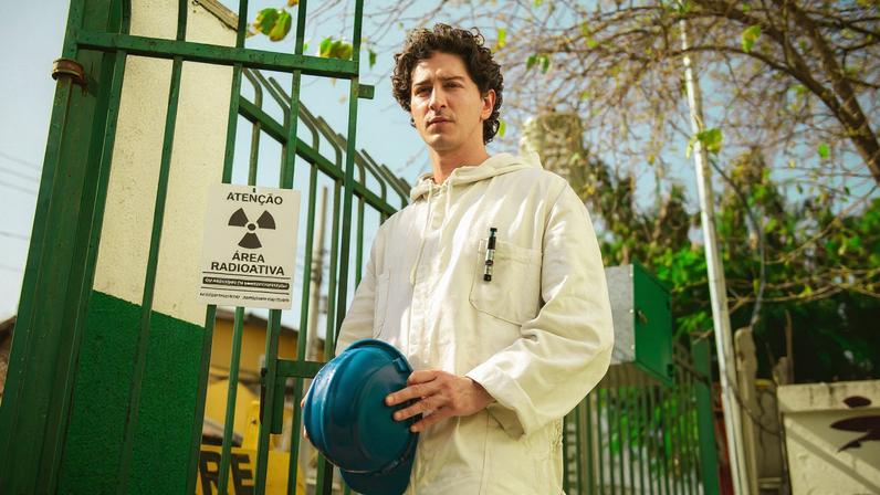 Homem jovem com macacão branco segura capacete azul em frente a portão verde com placa de aviso de área radioativa. Ambiente externo com árvores e céu claro ao fundo.