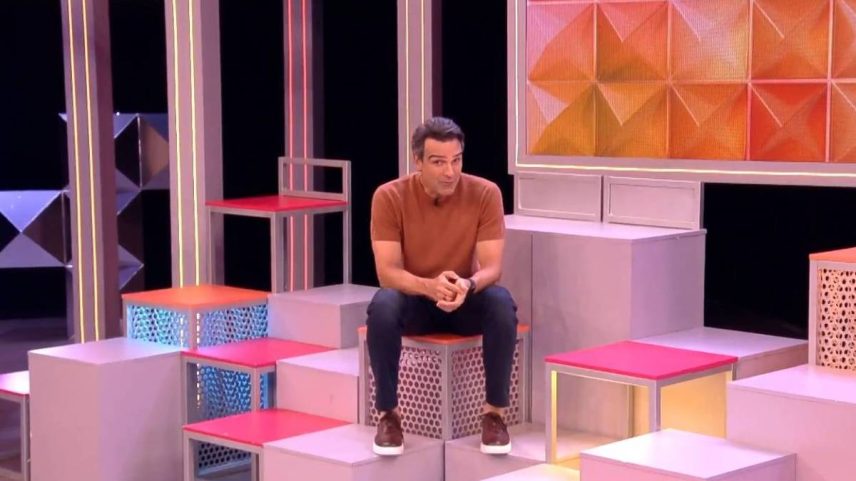 Homem de camiseta marrom e calça escura sentado em bloco branco com assento rosa em estúdio com blocos geométricos coloridos e painel laranja ao fundo.