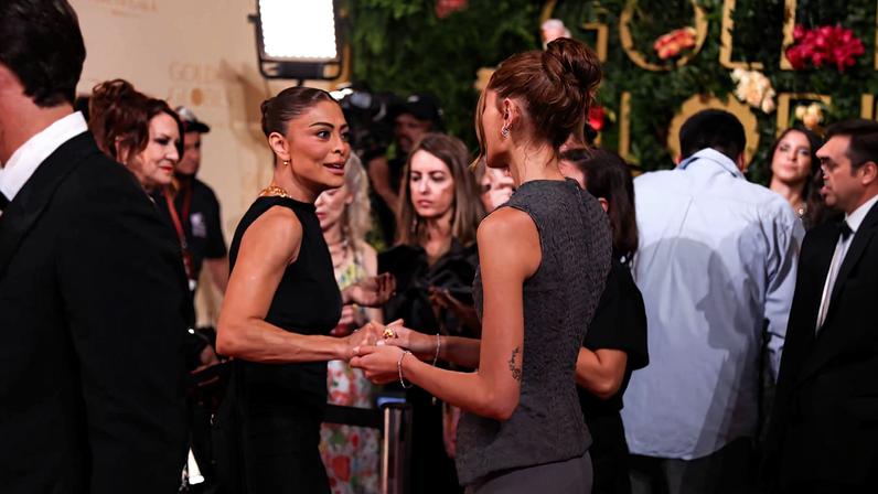 Duas mulheres de perfil conversam e seguram as mãos em evento social com várias pessoas ao redor. Uma veste vestido preto e a outra roupa cinza, com cabelo preso. Fundo com decoração verde e flores, iluminação artificial e outras pessoas observando.