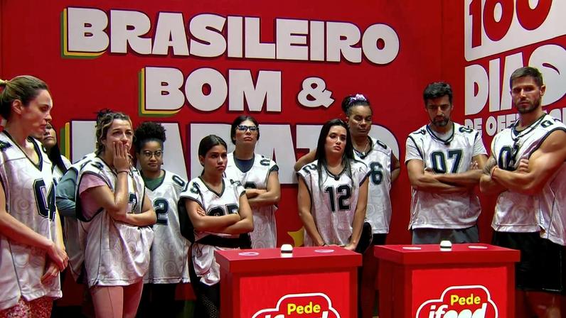 Dez pessoas vestindo coletes brancos numerados estão alinhadas em estúdio com paredes vermelhas. Ao fundo, texto grande diz 'BRASILEIRO BOM &'. Dois totens vermelhos com logo do iFood estão à frente do grupo.