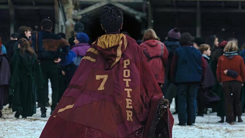 Pessoa de costas usando capa de quadribol vermelha com nome 'Potter' e número 7, caminhando em área com neve. Grupo de pessoas ao fundo em frente a estrutura de madeira com bandeiras coloridas.