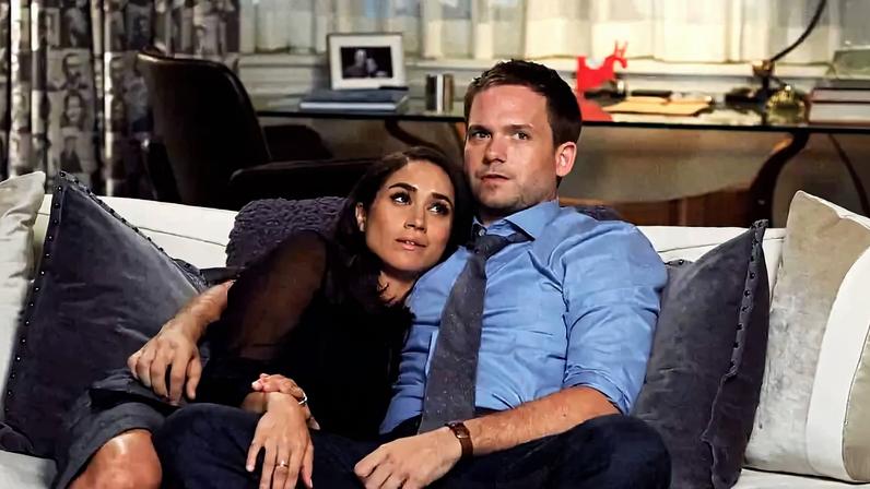 Homem de camisa azul e mulher de blusa preta sentados juntos no sofá, ela encostada nele. Ao fundo, mesa com livros, abajur e cortinas fechadas.