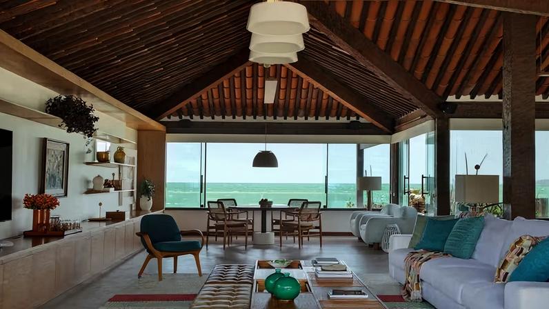 LIVING | Os janelões permitem a visão do mar. Ao fundo, mesa Pistão, de Arthur Casas; cadeiras Oscar, de Sergio Rodrigues; e par de poltronas Madame, de Ricardo Van Steen, todas adquiridas na loja Arquivo Contemporâneo, que também forneceu a poltrona Mia, de Jader Almeida, no tom de azul, voltada para a sala de estar. A luminária pendente Bossa, da Lumini foi instalada acima da mesa, enquanto o trio do modelo Nord, da mesma marca, fica no teto acima da mesa de centro Rosá, de Maria Cândida Machado. Nas prateleiras da estante, objetos da Ekko Home, Habito e Arquivo Contemporâneo, além de quadros do artista Marcelo Catalano